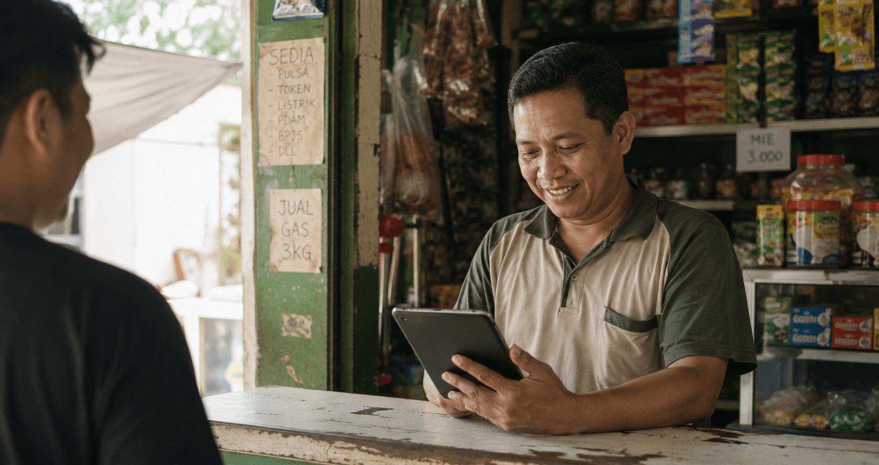 Pemilik warung tersenyum sambil mengecek tablet di meja lapaknya, dengan papan tulisan tangan "Sedia Pulsa, Token Listrik" di tiang warung.